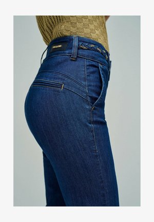 Persona con vaqueros ajustados azul oscuro, cinturón trenzado y camiseta de manga corta texturizada color marrón claro, vista lateral de la cintura al muslo.