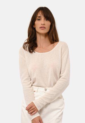 Beiger, langärmliger Strickpullover mit weitem Rundhalsausschnitt. Weiche Textur und eng anliegende Bündchen, kombiniert mit weißen hoch taillierten Hosen.