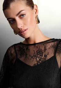 Haut en dentelle noire avec des motifs floraux par-dessus une robe noire, présentant un encolure large et de grandes boucles d'oreilles en or.