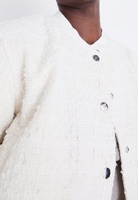 White textured jacket with a quilted pattern, featuring silver snap buttons and a ribbed collar. The fabric appears soft and structured.