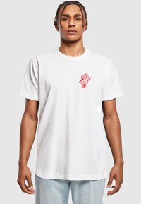 T-shirt en coton blanc avec un design de personnage de dessin animé rouge sur la poitrine gauche. Manches courtes et col rond. Coupe décontractée.