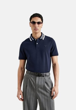 Uomo che indossa una polo blu navy con colletto a righe e scritte, pantaloni grigi plissettati, cintura nera, occhiali da sole e orecchini, in piedi su sfondo bianco.