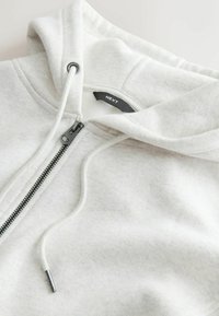 Sweat à capuche gris clair à fermeture éclair avec cordons et une étiquette noire visible à l'intérieur du col indiquant "NEXT".