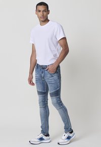 Jeans ajustados azules con acentos texturizados y rayas horizontales en las rodillas, combinados con una camiseta blanca de cuello redondo y calzado deportivo.