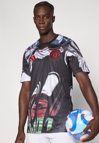 Maglia atletica nera con un design astratto multicolore, logo rosso sul petto, maniche corte e tessuto liscio e leggero. Una palla da calcio è tenuta in una mano.