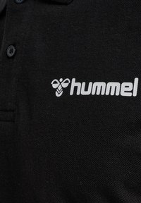 Svart pikétröja i texturerad tyg, med en vit "hummel"-logotyp och en bie-design, med tre knappar vid kragen.