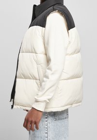 Puffweste mit schwarzem Oberteil und cremefarbenem Unterteil, mit hohem Kragen, seitlichem Reißverschluss und seitlichen Taschen. Strukturiertem Stoff mit gesteppte Details.