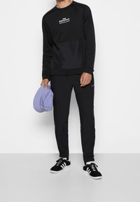 Svart långärmad sweatshirt med en lila panel och logo, tillsammans med svarta byxor och svarta skor. Håller en ljuslila hatt.