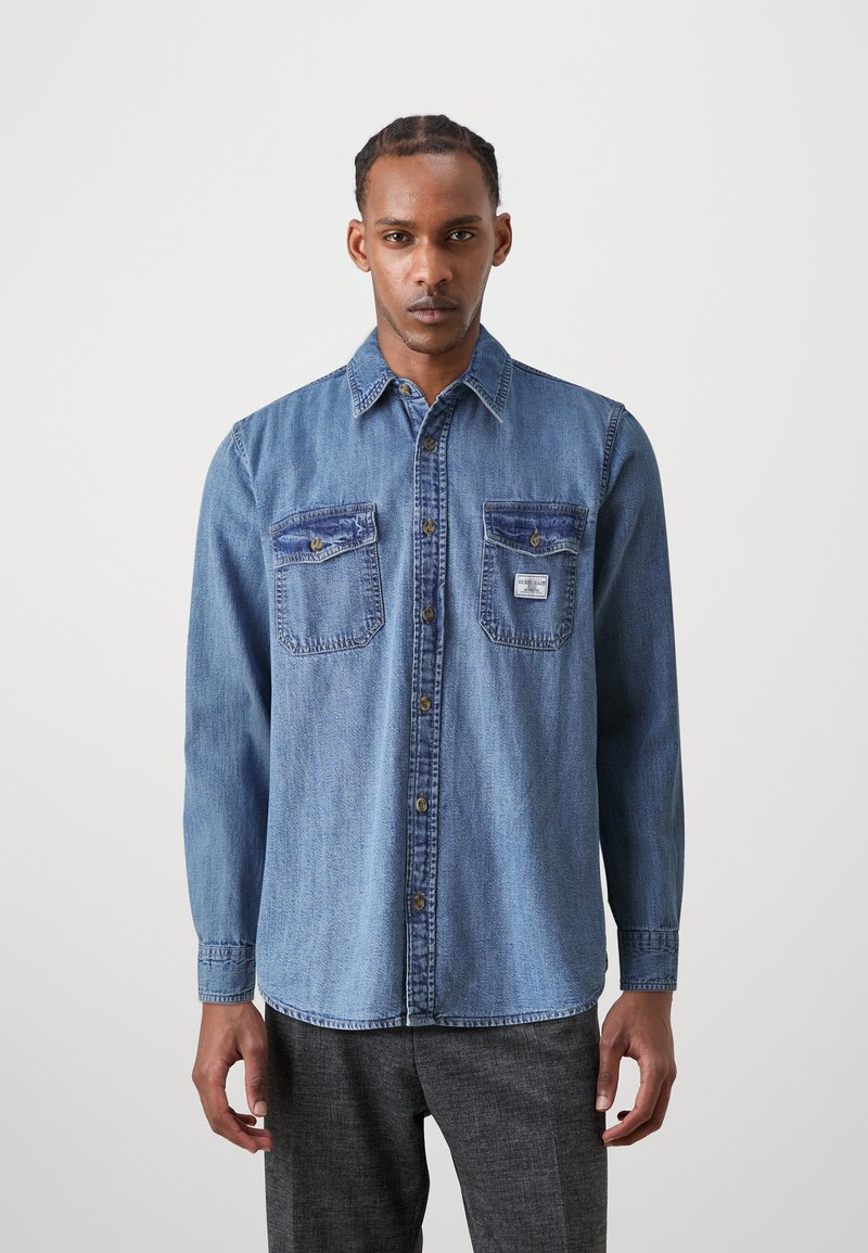Hellblaue Denim-Hemdjacke mit Knopfleiste, zwei Fronttaschen, kontrastierenden Nähten und einem Kragen. Lange Ärmel mit Knopfverschlüssen.