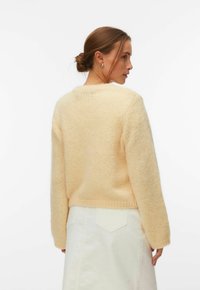 Femme aux cheveux bruns attachés en chignon, portant un pull en laine crème et une jupe blanche, vue de dos sur un fond uni.
