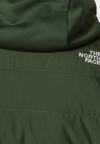 Veste matelassée vert foncé avec capuche, affichant le logo « The North Face » brodé en blanc sur la partie supérieure droite du dos.