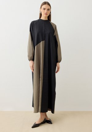 Kvinde står iført en lang sort og beige farveblokkjole med lange ærmer og sorte spidse flade sko mod en ensfarvet hvid baggrund.