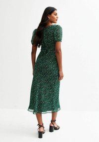 Robe midi florale verte avec de courtes manches bouffantes, en tissu doux, taille cintrée et jupe fluide. Des sandales à talons noires complètent la tenue.