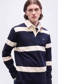 Νεαρός άνδρας με μεσαίου μήκους μαλλιά, φοράει ριγέ μπλουζάκι ράγκμπι σε ναυτικό μπλε και μπεζ χρώμα, πάνω από ένα πουκάμισο με γιακά και γραβάτα, στέκεται μπροστά σε λευκό φόντο.