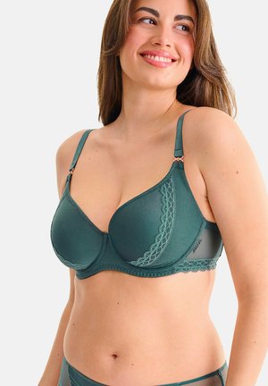 Femme aux longs cheveux bruns souriante, portant un soutien-gorge vert foncé orné de dentelle et des sous-vêtements assortis sur un fond uni clair.