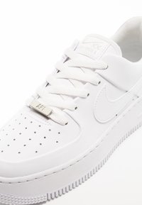 Białe sneakersy Nike Air Force 1 z białymi sznurówkami, perforowaną częścią palców, metalowym znaczkiem na sznurówkach oraz haftowanym logo Nike w kształcie swoosha.