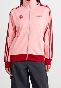 Světle růžová bunda na zip s červenými akcenty, prezentující tři červené pruhy na rukávech a vyšívané logo na hrudi. Hladká látka.