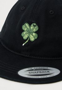 Black snapback cap with embroidered green four-leaf clover on front and a circular brand sticker on the brim.