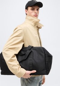 Νεαρός άνδρας με μπεζ μπουφάν και μαύρο καπέλο, κρατώντας μια μεγάλη μαύρη τσάντα ταξιδιού μπροστά σε απλό φωτεινό φόντο.