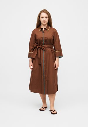 Ženska stoji v rjavi midi obleki z zlatim okrasjem, zavezanim pasom v obliki pentlje in črnimi japonkami na belem ozadju.