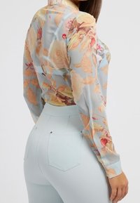 Blouse fleurie transparente à manches longues en bleu pastel, ornée d'imprimés de roses roses et jaunes, associée à un pantalon taille haute bleu clair.