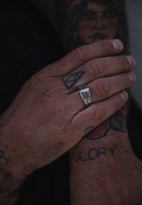 Sidabrinis žiedas su raižytu ornamentu, esantis ant rankos, kuri puikuojasi tatuiruotėmis. Ant pirštų matyti tekstūruota oda ir sudėtingi raštai.