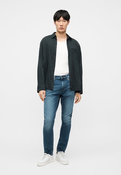 Νεαρός άνδρας που στέκεται, φοράει σκούρο ανοιχτό πουκάμισο, λευκό φανελάκι, μπλε τζιν και λευκά αθλητικά παπούτσια, μπροστά σε ένα απλό φωτεινό φόντο.