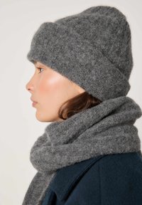 Profiilvaates naine, kes kannab paksu halli kootud mütsi ja sobivat salli tumeda välisriietuse vastu lihtsa taustaga.