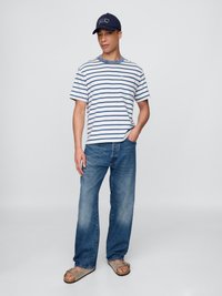 Stripet t-skjorte i marineblå og hvit, avslappet passform; kombinert med lyseblå denimjeans; brune sandaler; marineblå caps med hvit tekstdetalj.