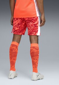 Maillot de sport orange associé à un short à motifs rouges, des chaussettes orange jusqu'aux genoux et des chaussures blanches. Design graphique distinctif sur le short et les chaussettes.