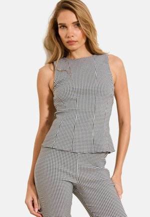 Fekete-fehér kockás ujjatlan felsőt és hozzáillő, testhezálló nadrágot viselő nő, egy egyszínű, világos háttér előtt áll.