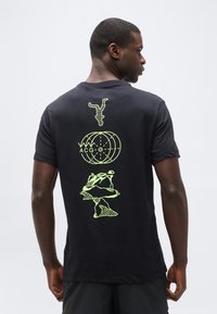 Νέος άνδρας με μαύρο μπλουζάκι που έχει τυπωμένες στο πίσω μέρος νέον κίτρινες αφηρημένες φιγούρες αστροναύτη, υδρογείου και πετρωμάτων, στέκεται μπροστά σε μια απλή φόντο.