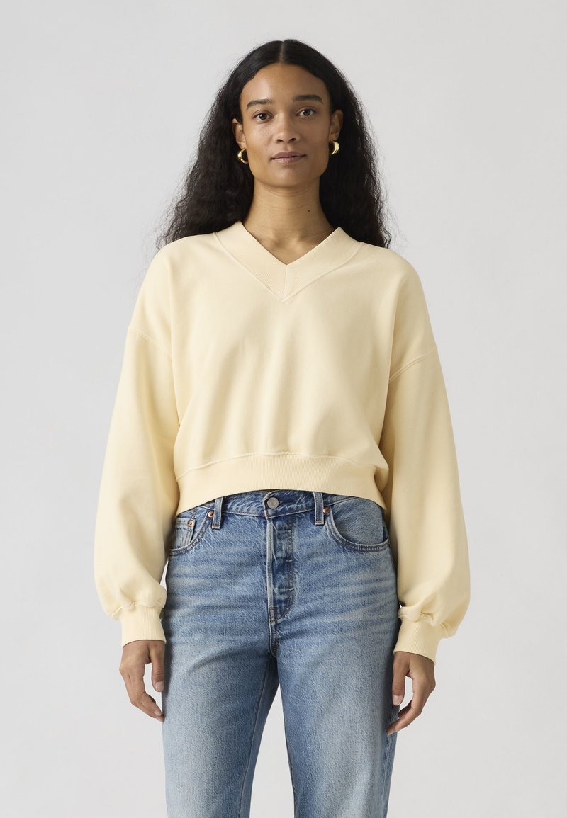 Femme aux longs cheveux bouclés et foncés, portant des boucles d'oreilles créoles dorées, un sweat-shirt crème à col en V et un jean taille haute bleu clair, debout devant un fond uni.