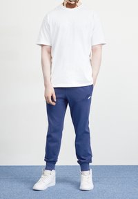 Bílé bavlněné tričko s malým logem, kombinované s modrými sportovními kalhotami a bílými teniskami. Oblečení má volný střih a jednoduchý design.