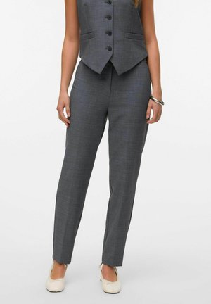 Gilet grigio su misura con cinque bottoni neri, abbinato a pantaloni skinny coordinati. Il materiale sembra avere una texture. Gli accessori includono un bracciale d'argento.