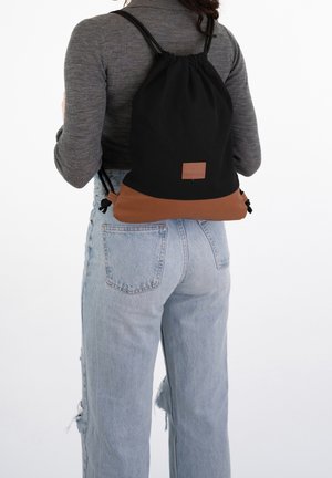 Une personne portant un jean déchiré bleu clair et un haut côtelé gris porte un sac à dos à cordon noir et marron dans le dos.