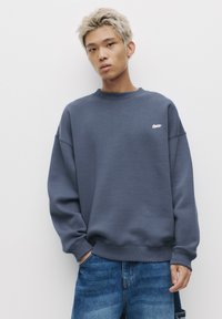 Sötétkék pulcsi, puha anyagból készült. Kerek nyakú, esett vállú, és egy kis fehér logó található a mellkason.