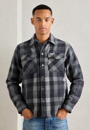 Hombre vistiendo una camisa de cuadros gris y negra con botones a presión y bolsillos en el pecho, con las manos en los bolsillos de unos jeans azules, fondo neutro.
