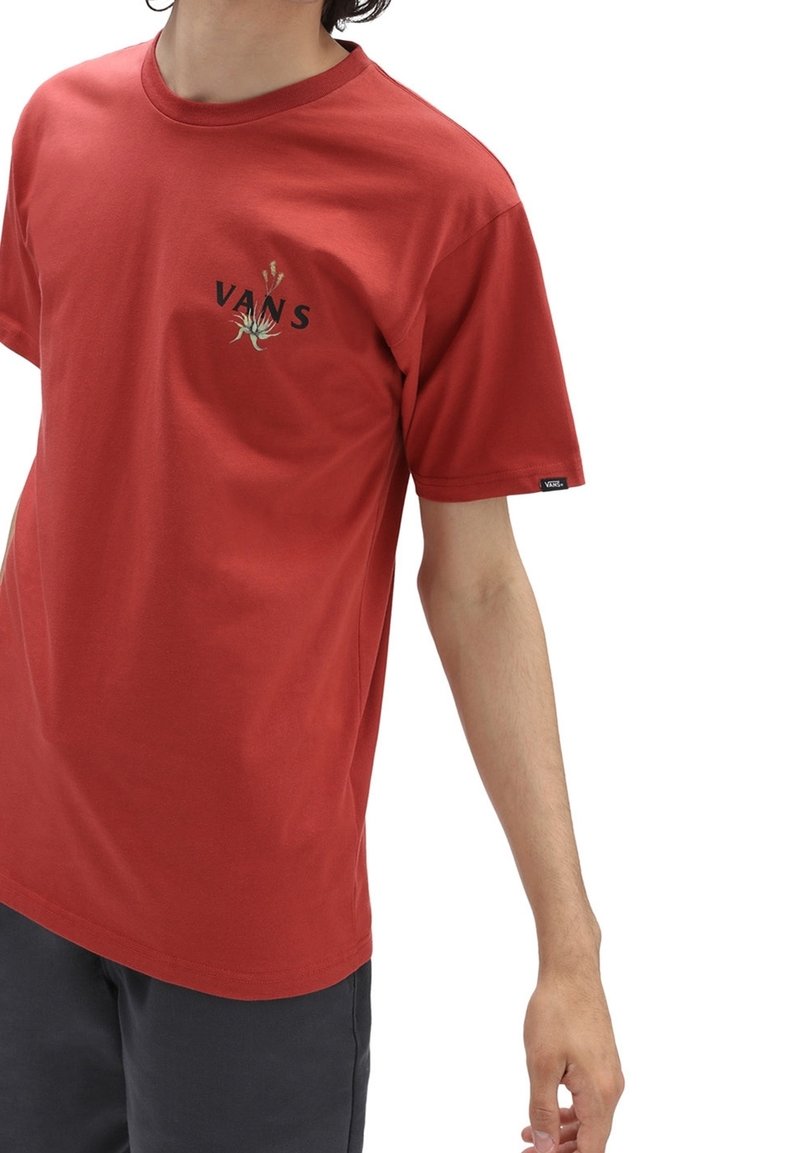Personne portant un t-shirt Vans couleur rouille avec un petit graphique de plante et un logo sur la poitrine, associé à un pantalon foncé.