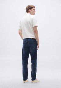 Bijela polo majica, plave traperice i svijetle tenisice. Traperice imaju kožni zakrpu i uskog su kroja. Pozadina je jednostavna i svijetla.