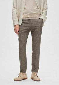 Chemise boutonnée en lin beige clair sur un pull neutre, associée à un pantalon marron texturé et des chaussures claires. Éléments de design simples et modernes.
