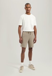 Hvit bomull t-skjorte med lomme, kombinert med lysebrune teksturerte shorts og beige joggesko. Modellen står foran en nøytral bakgrunn.