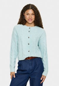 Vrouw met krullend bruin haar draagt een lichtblauwe kabelgebreide cardigan met zilveren knopen en hooggesneden donkerblauwe spijkerbroek met een bruine riem.
