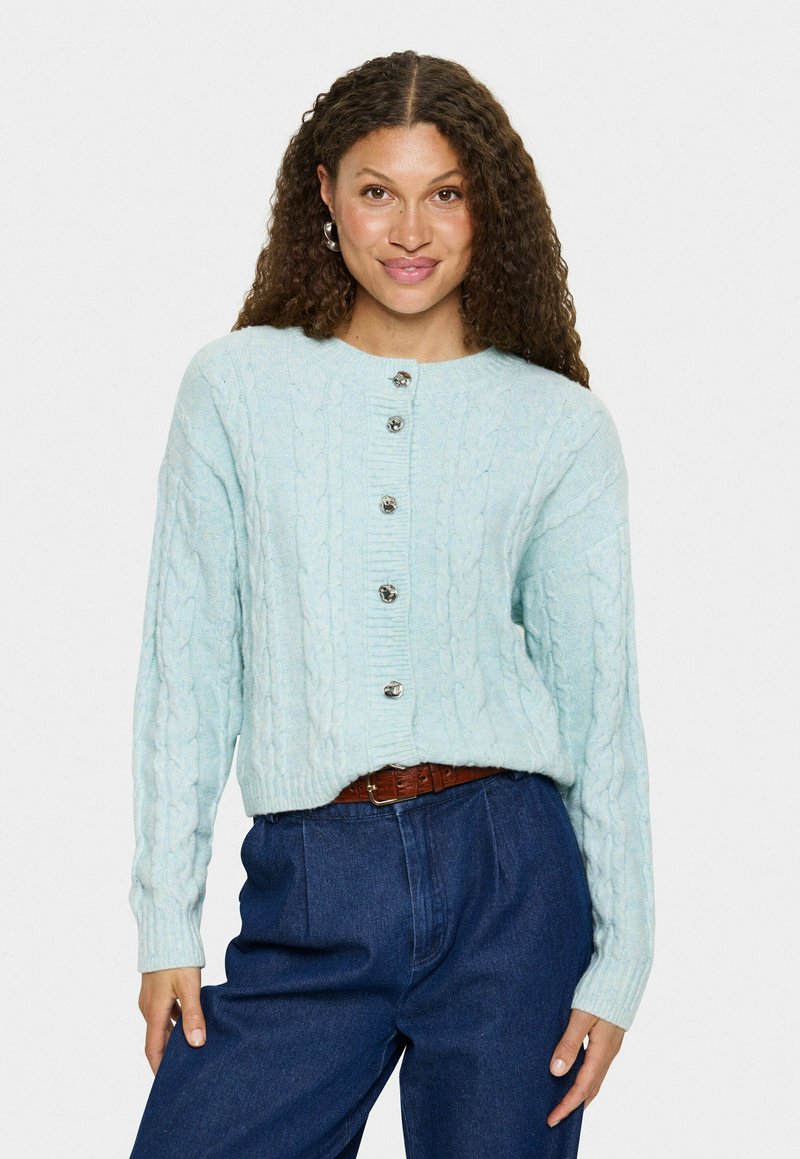Vrouw met krullend bruin haar draagt een lichtblauwe kabelgebreide cardigan met zilveren knopen en hooggesneden donkerblauwe spijkerbroek met een bruine riem.