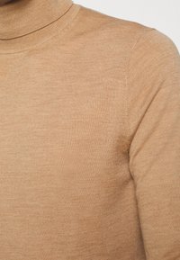 Pull à col roulé beige en tricot doux et extensible ; coupe ajustée avec texture côtelée et encolure propre et sans couture.