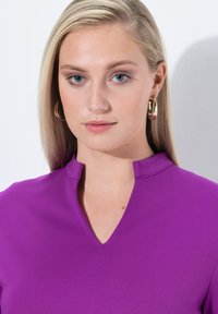 Haut en V en tissu violet vif avec une texture lisse. Le modèle porte de grandes boucles d'oreilles en or. L'arrière-plan est blanc pour le contraste.