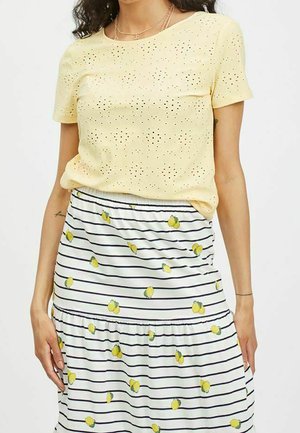 Kvinde iført en lysegul øjlet kortærmet top og et hvidt skørt med sorte striber og spredte citrontryk.