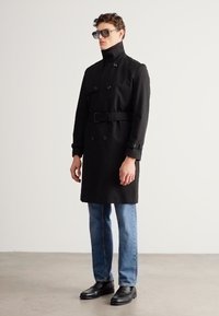 Mustast topeltkarvaga trentsmantel kõrge kraega, vööga taljes ja varrukate detailidega, kombineeritud helesiniste teksade ja mustade kingadega.