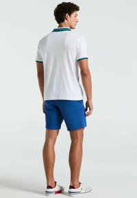 Weißes Polohemd mit türkisfarbenen und dunkelblauen Akzenten, kombiniert mit blauen Shorts. Model trägt weiße Sneakers mit marineblauen und roten Details.