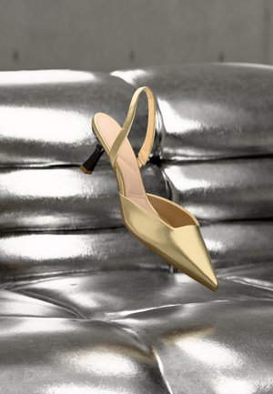 Pantof cu toc înalt, vârf ascuțit și bretea slingback, plutind deasupra unei canapele din piele argintie metalizată, cu tapițerie capitonată.